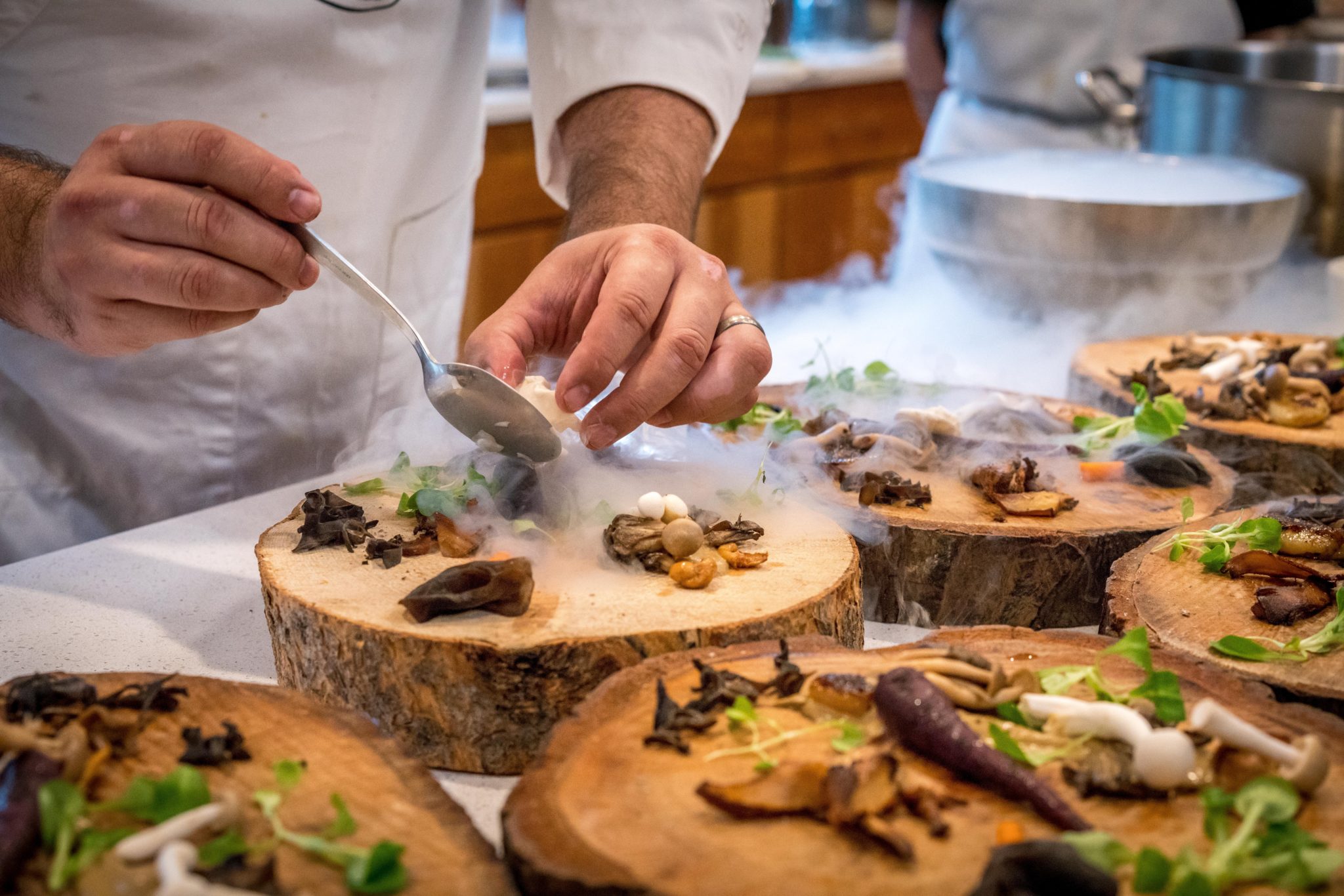 Chef meticulously garnishing mushroom dishes with fresh herbs on rustic wooden slabs amid a cloud of aromatic smoke in a professional kitchen.